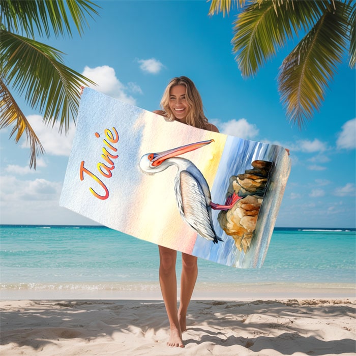 Woman holding a Personalized beach towel with a pelican design and 'Janie' text on a beach with palm trees and ocean in the background.