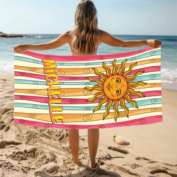 Person holding a colorful beach towel with a sun design and the name 'Michelle' on a sandy beach.